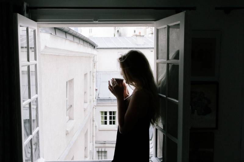 Silhouet van een vrouw met een kop koffie voor een raam, ochtendlicht over de stad