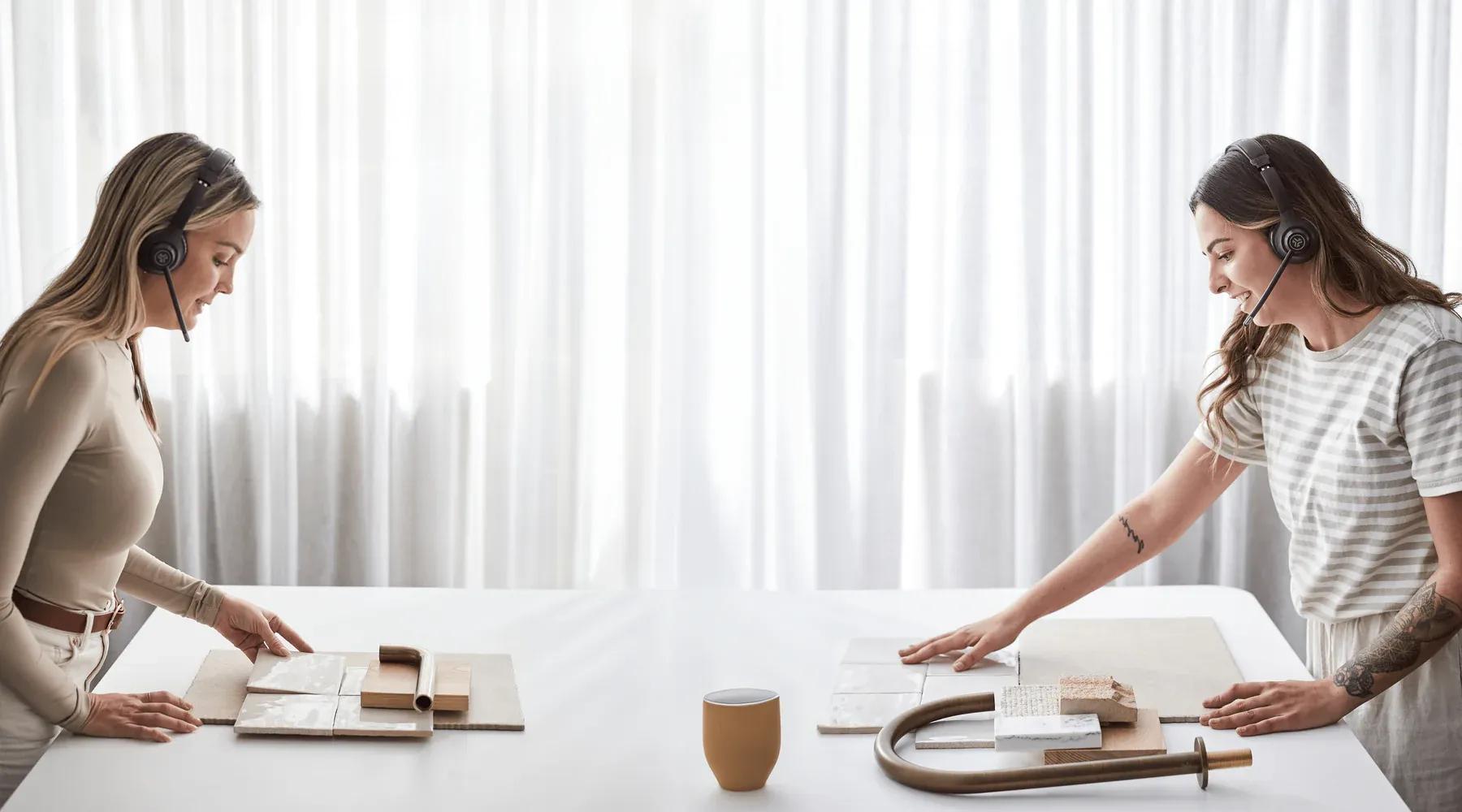 Two women wearing headsets compare tile and tapware samples across a table
