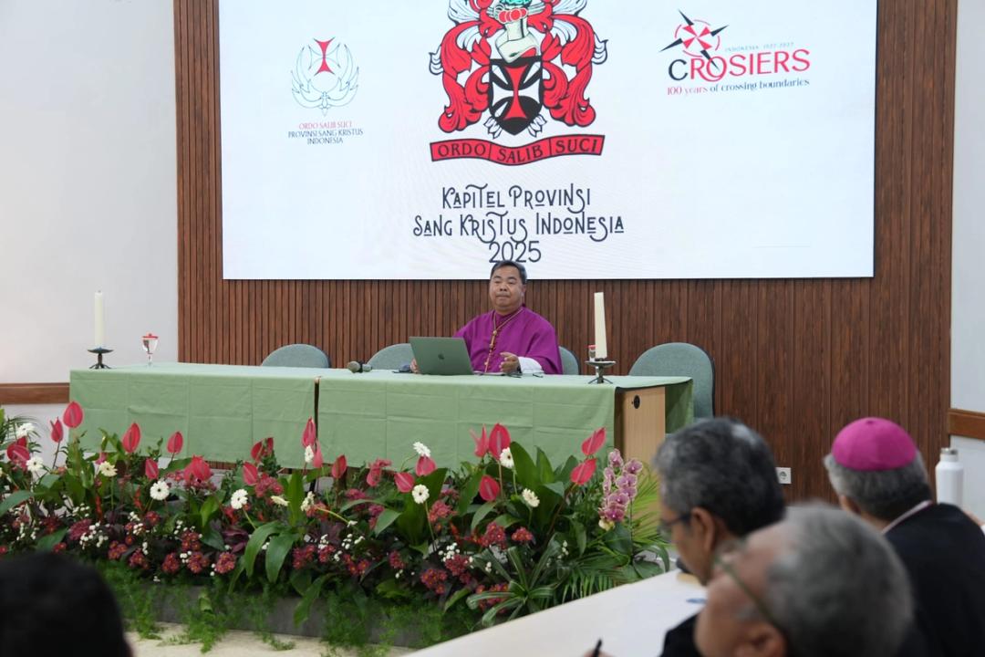 Seorang uskup berjubah ungu duduk di meja pimpinan di bawah layar bertuliskan Kapitel Provinsi Sang Kristus Indonesia 2025.