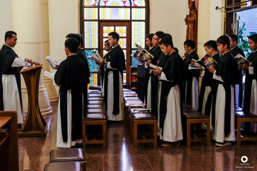 Para frater berjubah hitam-putih berdiri di bangku koor sambil bernyanyi dari buku doa.