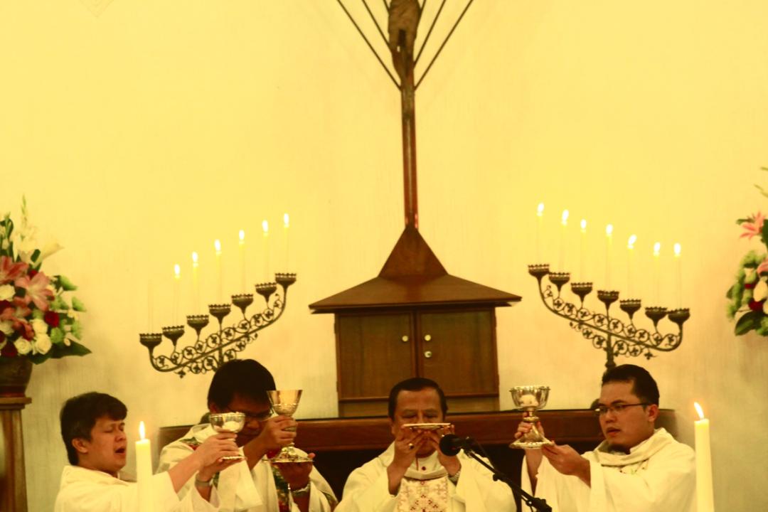 Empat imam berbusana liturgi putih mengangkat piala dan patena di altar, diapit kandelar dan bunga.