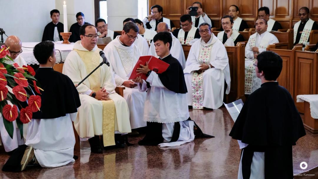 Seorang frater berlutut membacakan rumusan kaul dari buku merah di hadapan imam, disaksikan para Krosier.