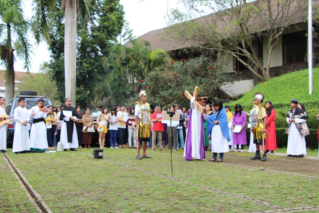 Drama Jalan Salib di halaman: pemeran Yesus memanggul salib kayu, diapit pemeran prajurit, ditonton umat.