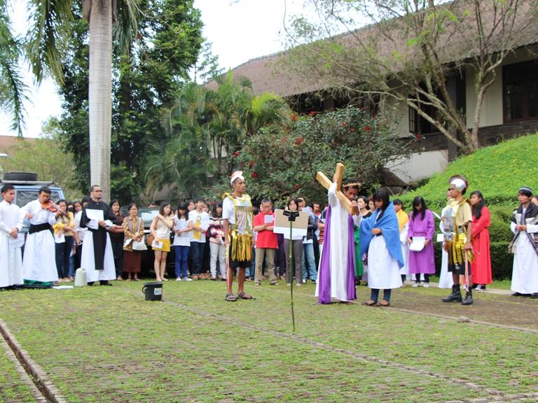 Drama Jalan Salib di halaman: pemeran Yesus memanggul salib kayu, diapit pemeran prajurit, ditonton umat.