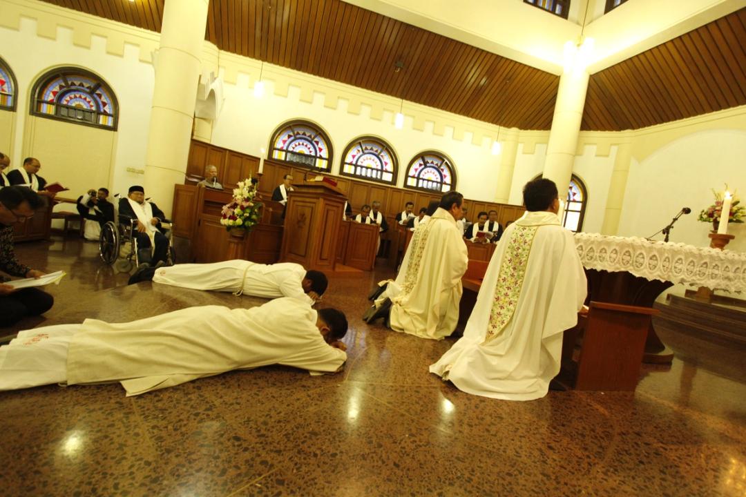 Dua pria berjubah putih bersujud tertelungkup di lantai gereja, sementara dua imam berkasula berlutut menghadap altar.