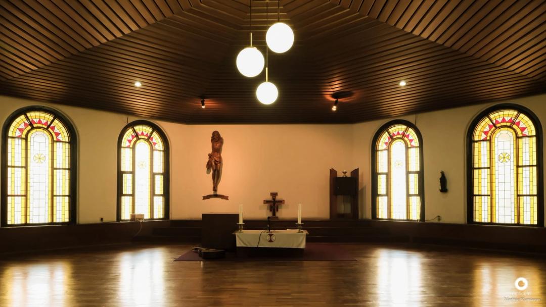 Kapel berlantai kayu dan berlangit-langit kayu dengan jendela kaca patri melengkung dan salib tergantung di atas altar.