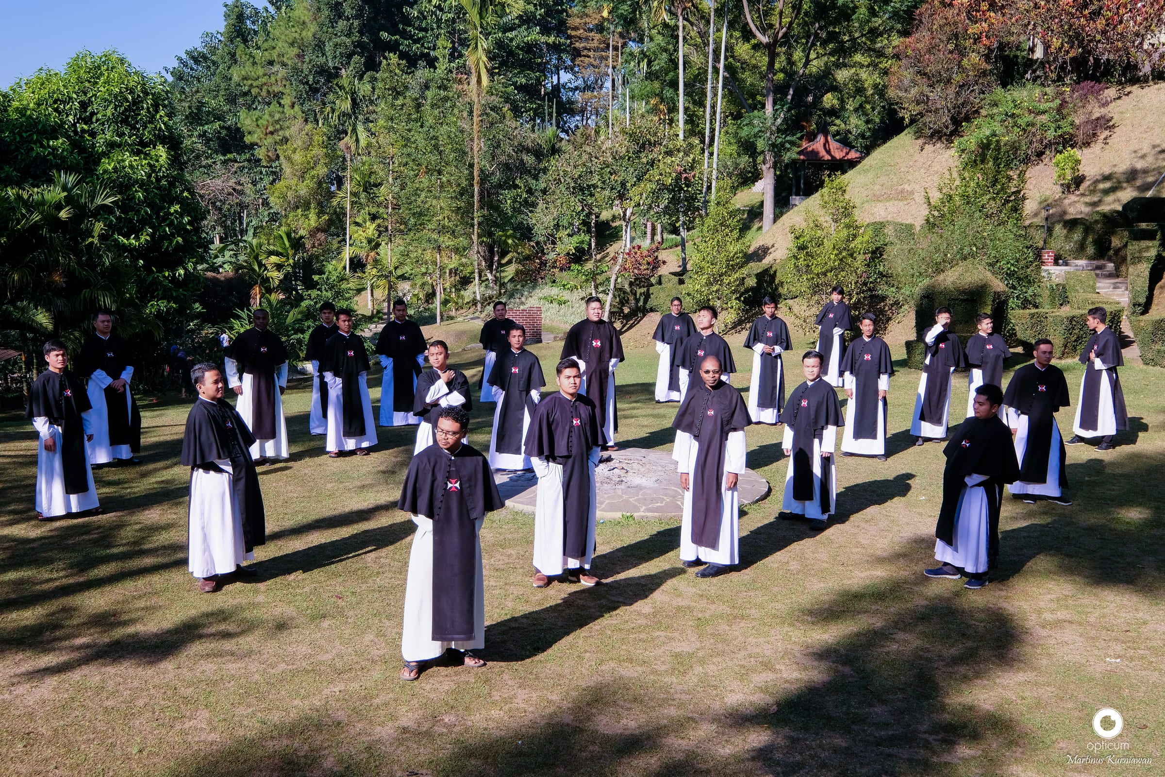 Para frater dan novis berjubah hitam-putih berdiri tersebar di lapangan rumput berlatar pepohonan.