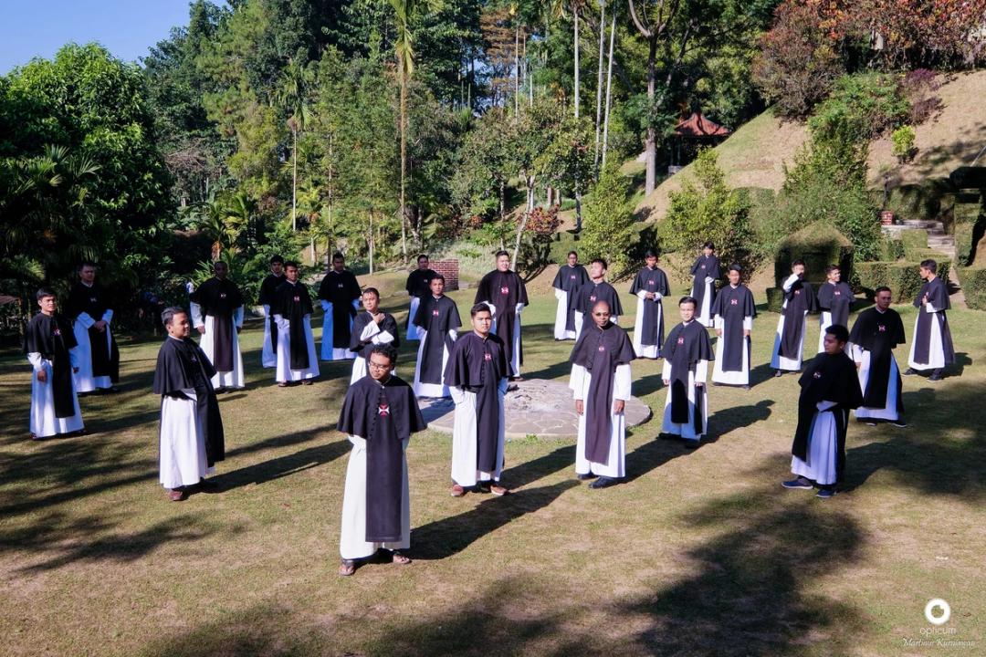 Para frater dan novis berjubah hitam-putih berdiri tersebar di lapangan rumput berlatar pepohonan.