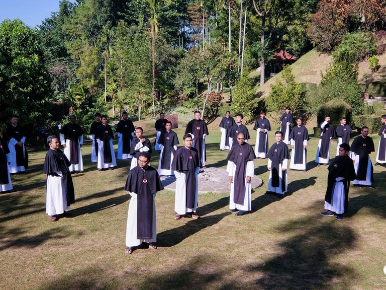 Para frater dan novis berjubah hitam-putih berdiri tersebar di lapangan rumput berlatar pepohonan.