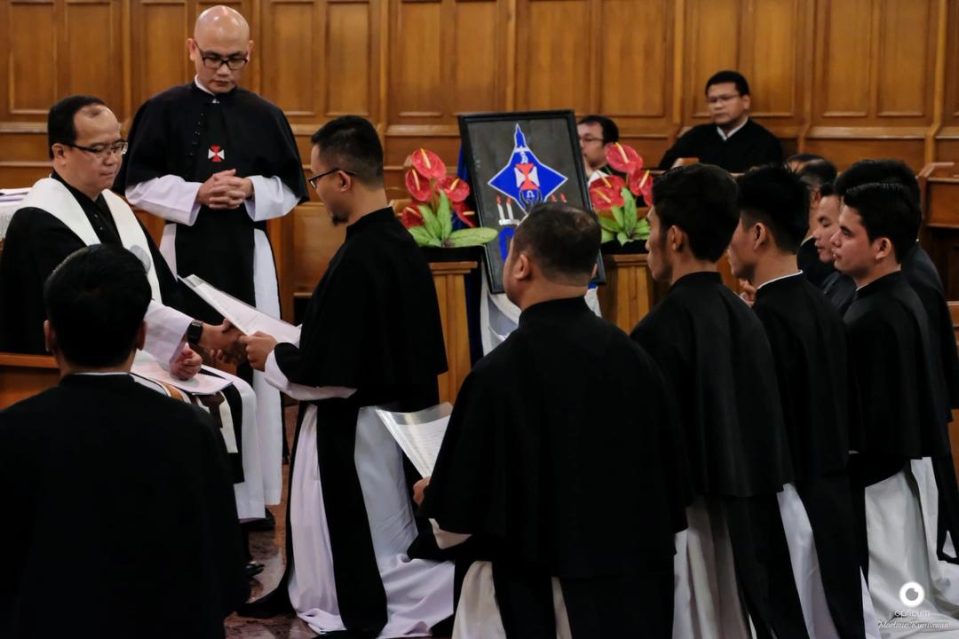 Para frater berjubah hitam-putih membaca lembaran di depan lambang Ordo yang dibingkai dan rangkaian bunga merah.