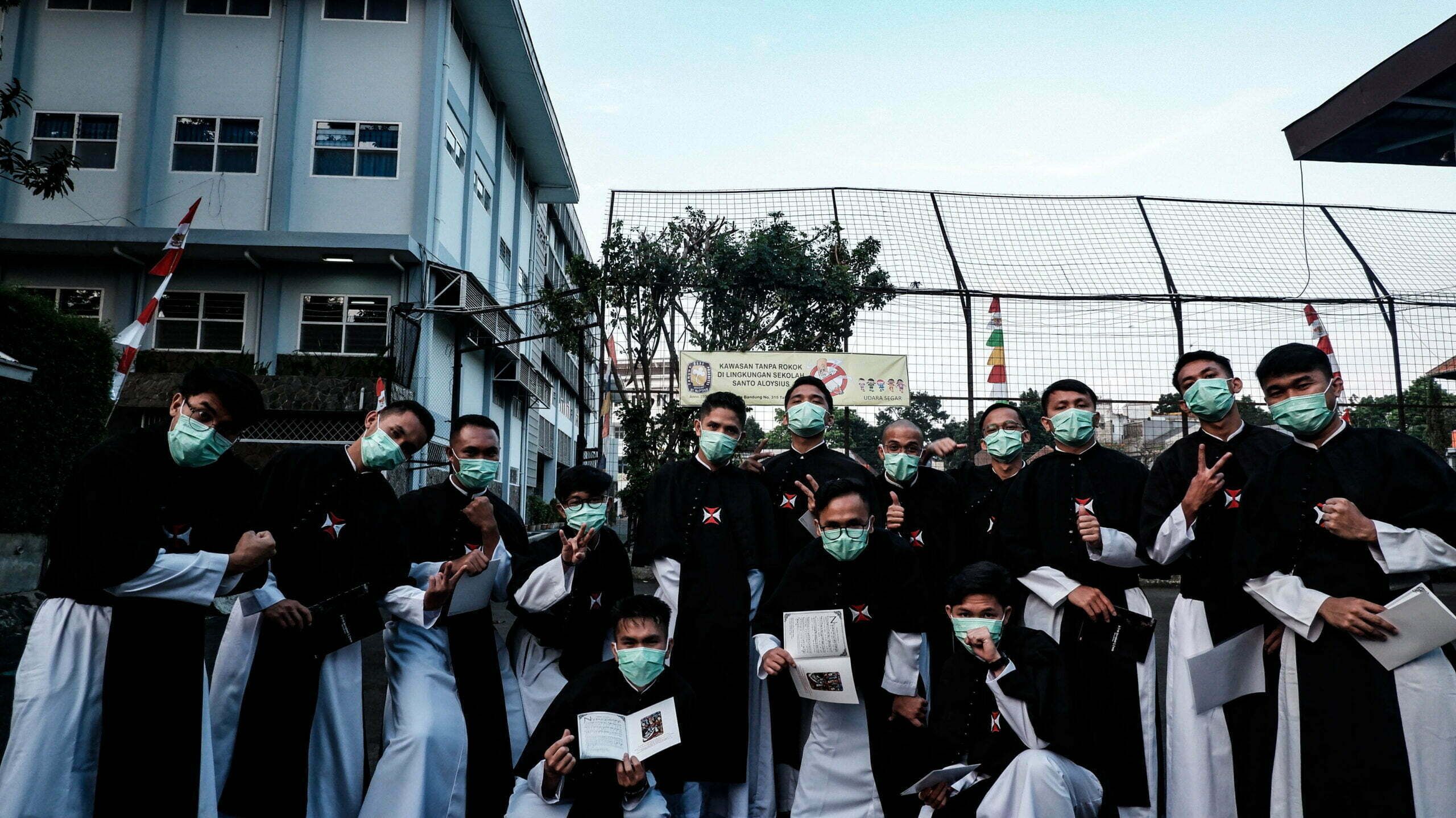 Para frater Krosier bermasker berpose bersama sambil memegang buku kecil di halaman sebuah sekolah.