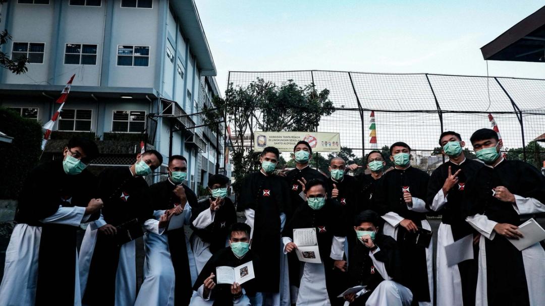Para frater Krosier bermasker berpose bersama sambil memegang buku kecil di halaman sebuah sekolah.