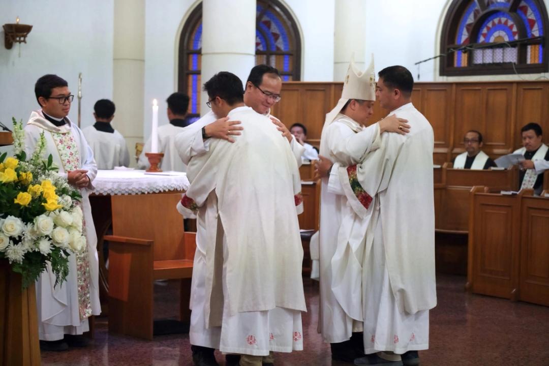 Para imam berbusana putih saling berpelukan di depan altar, disaksikan uskup dan para Krosier di bangku koor.