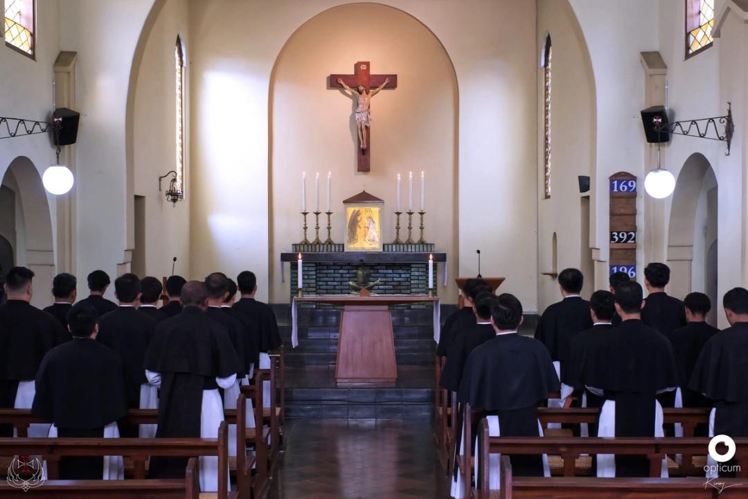 Barisan frater berjubah hitam-putih berdiri menghadap altar kapel yang diterangi lilin.