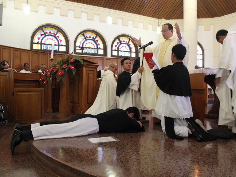 Seorang frater berjubah Krosier bersujud tertelungkup di depan altar, sementara seorang imam merentangkan tangan.