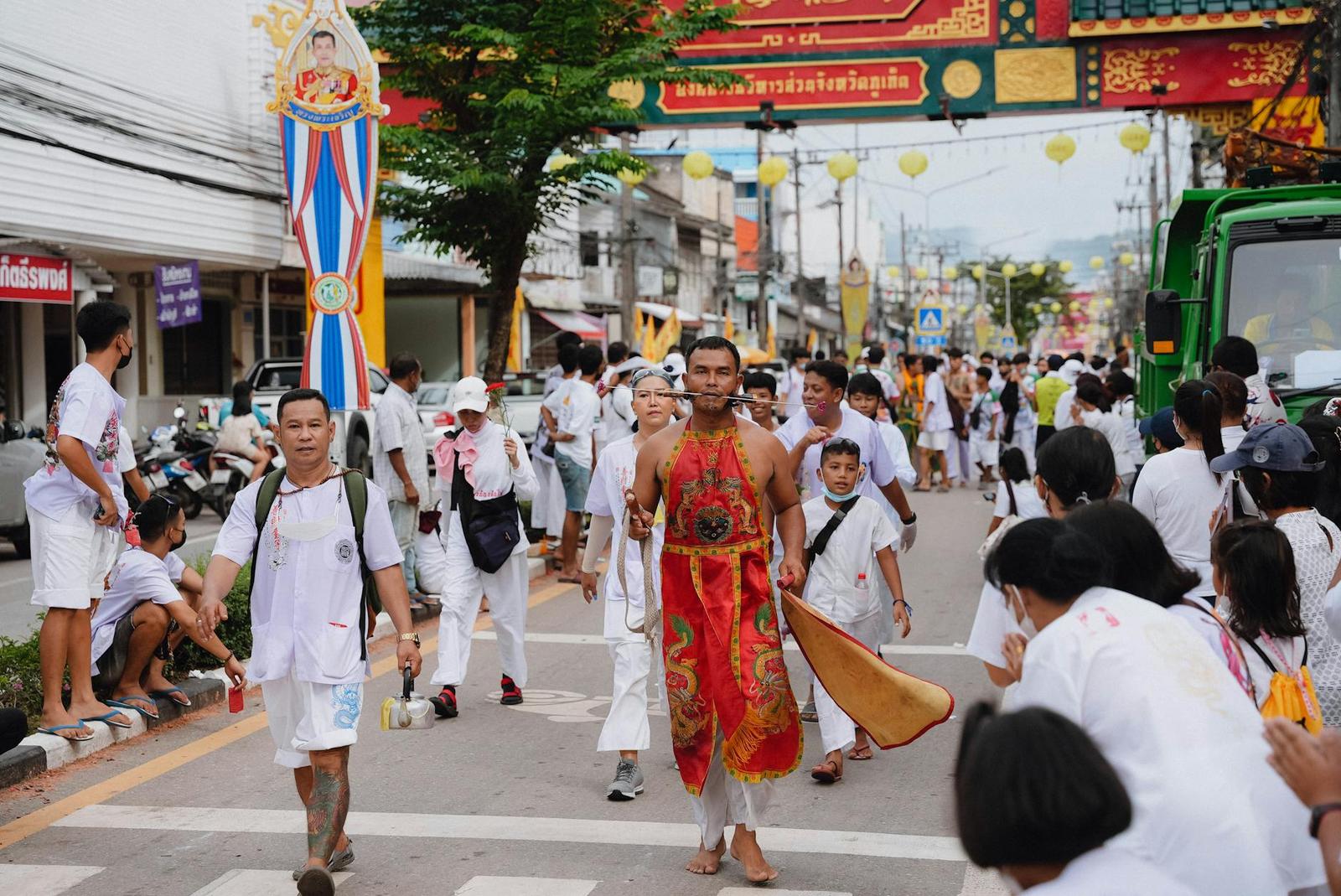 A street procession with a man in an ornate red costume leading a crowd dressed in white