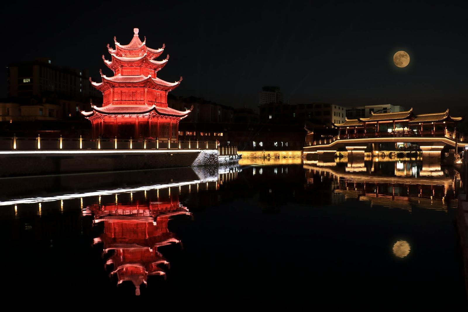 A red pagoda glowing at night beside still water under a full moon