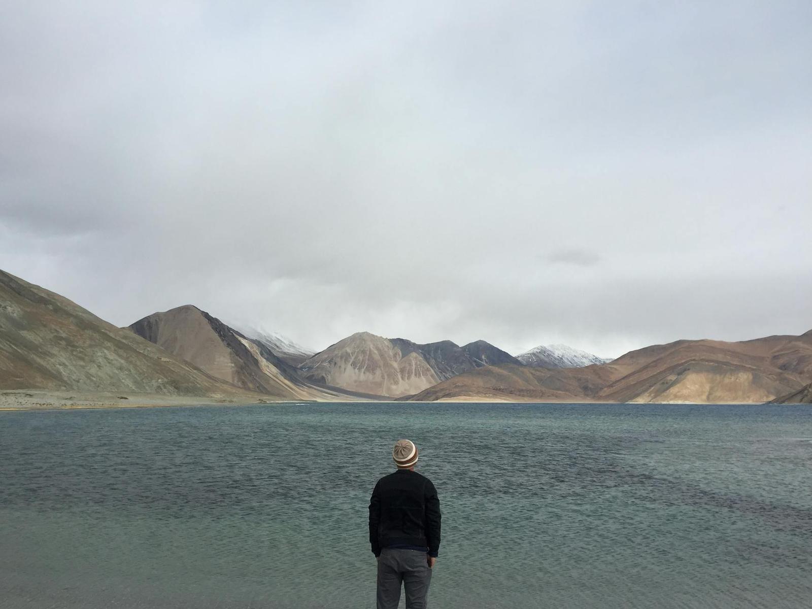 A person standing before a turquoise mountain lake under overcast skies