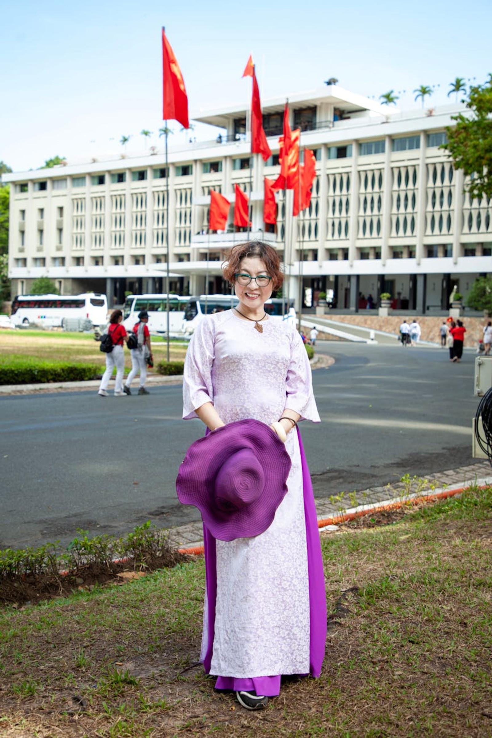 Woman in a lavender ao dai posing with a purple hat before a government building
