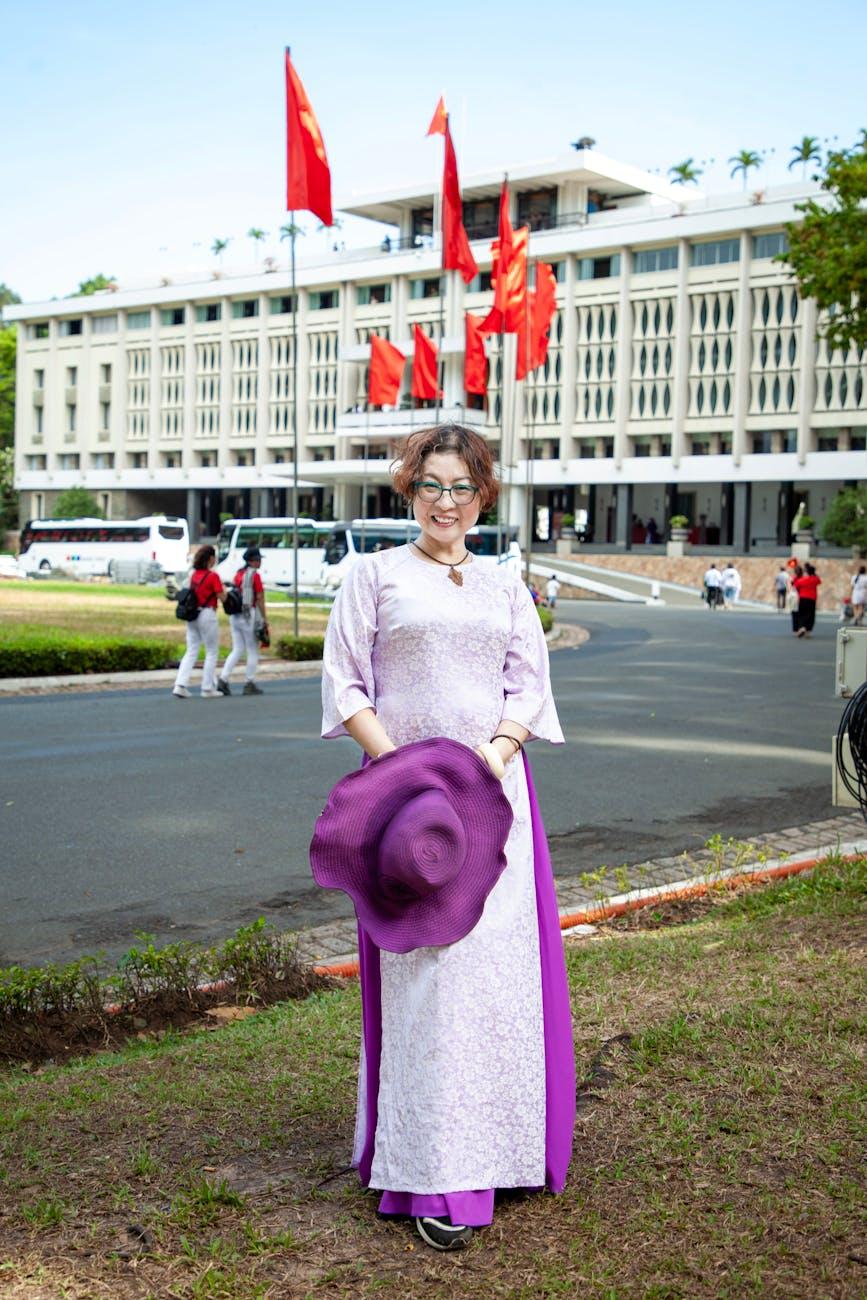 Woman in a lavender ao dai posing with a purple hat before a government building