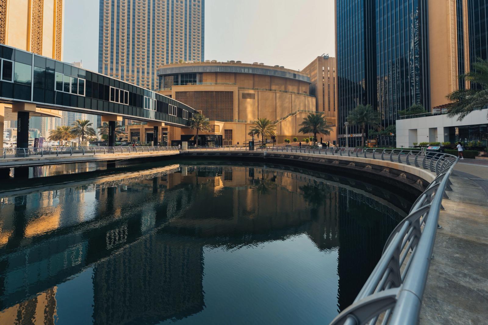 A curved waterfront promenade beside glass towers reflected in a still canal