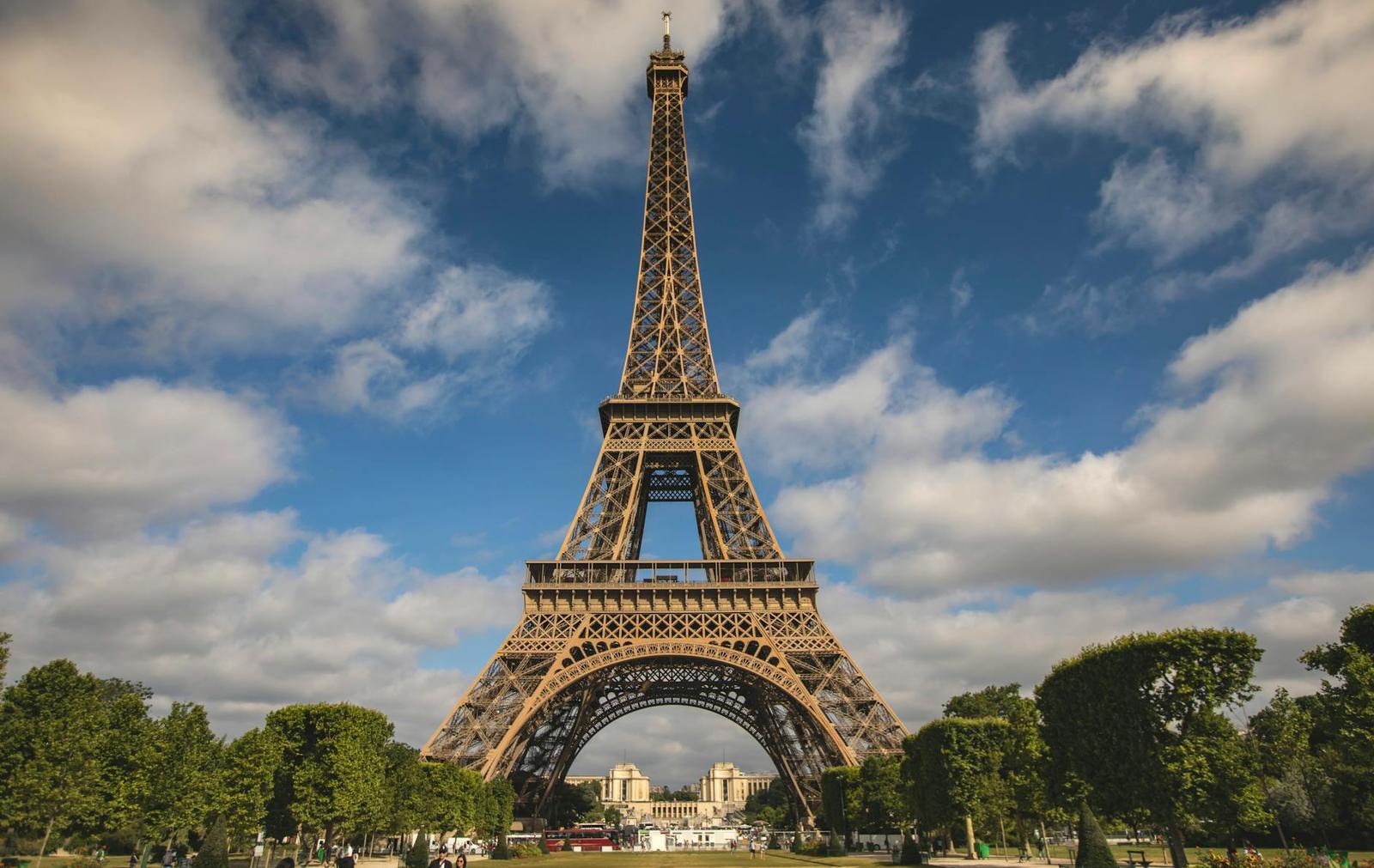 The Eiffel Tower rising against a partly cloudy blue sky above tree-lined gardens