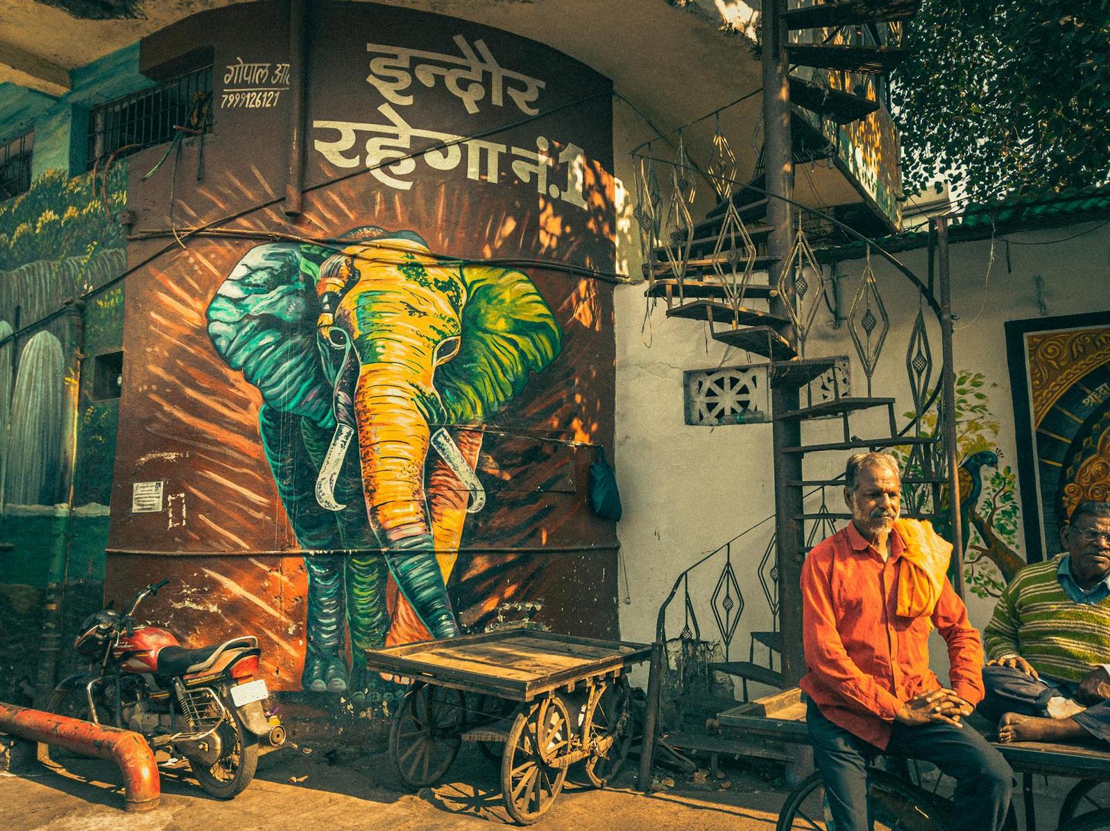 A colourful elephant mural above two men seated beside a parked motorcycle