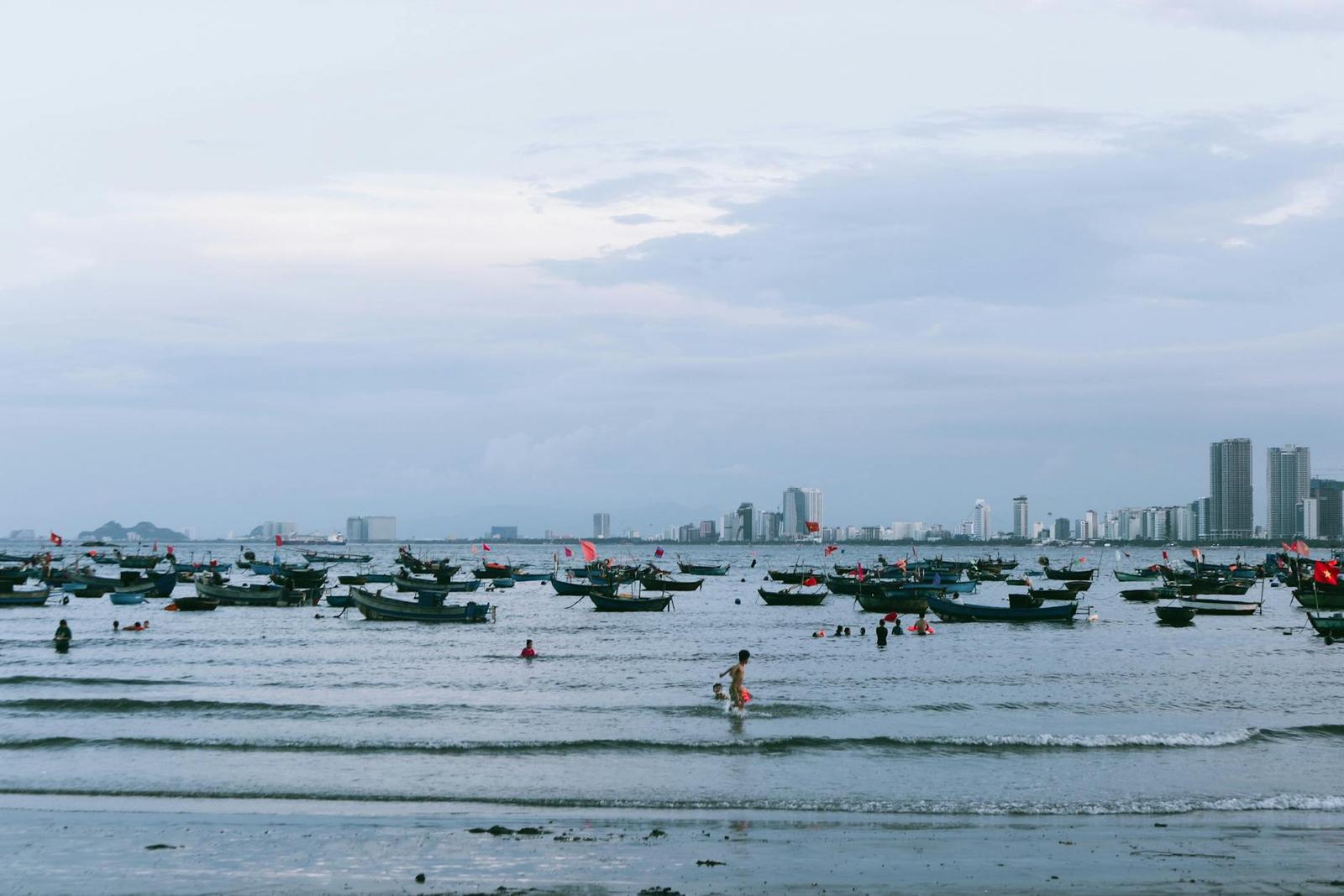 Fishing boats anchored off a beach with people wading in the shallows and a city skyline behind