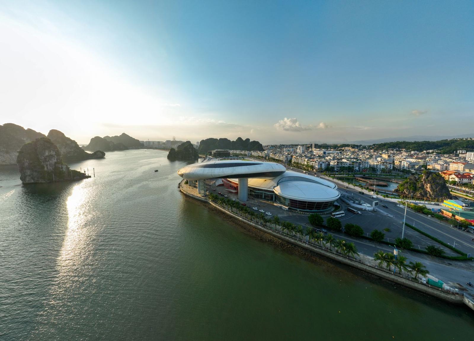 An aerial view of a curved waterfront building beside limestone islands and a bay at sunset