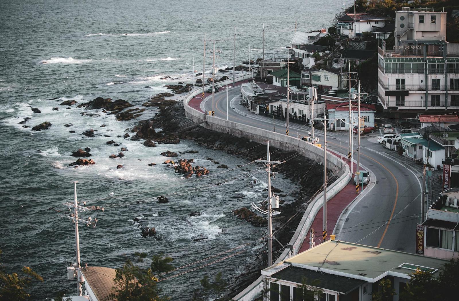 A coastal road curves along a rocky shoreline lined with hillside buildings