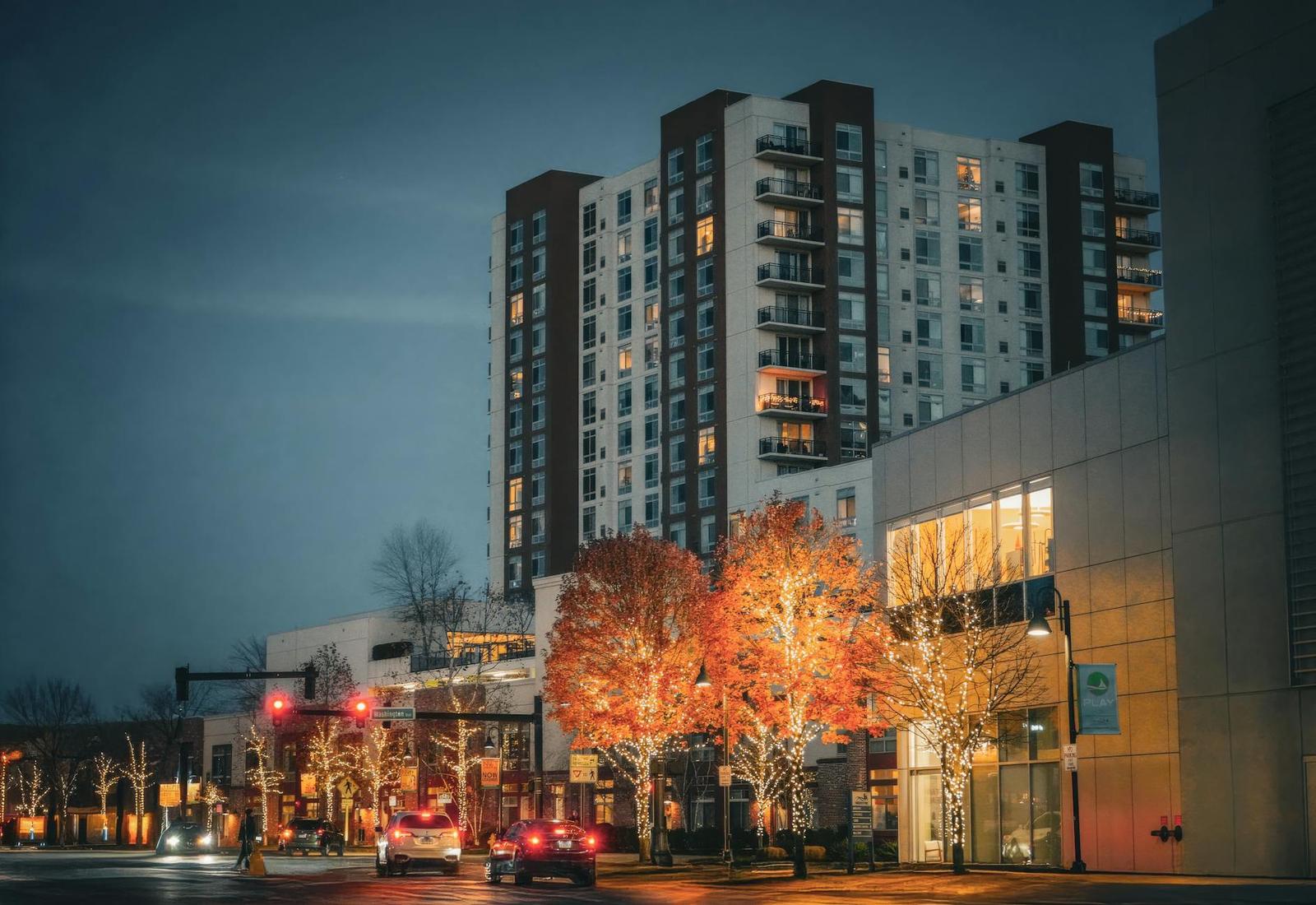 A high-rise apartment building at night beside a street lined with lit autumn trees