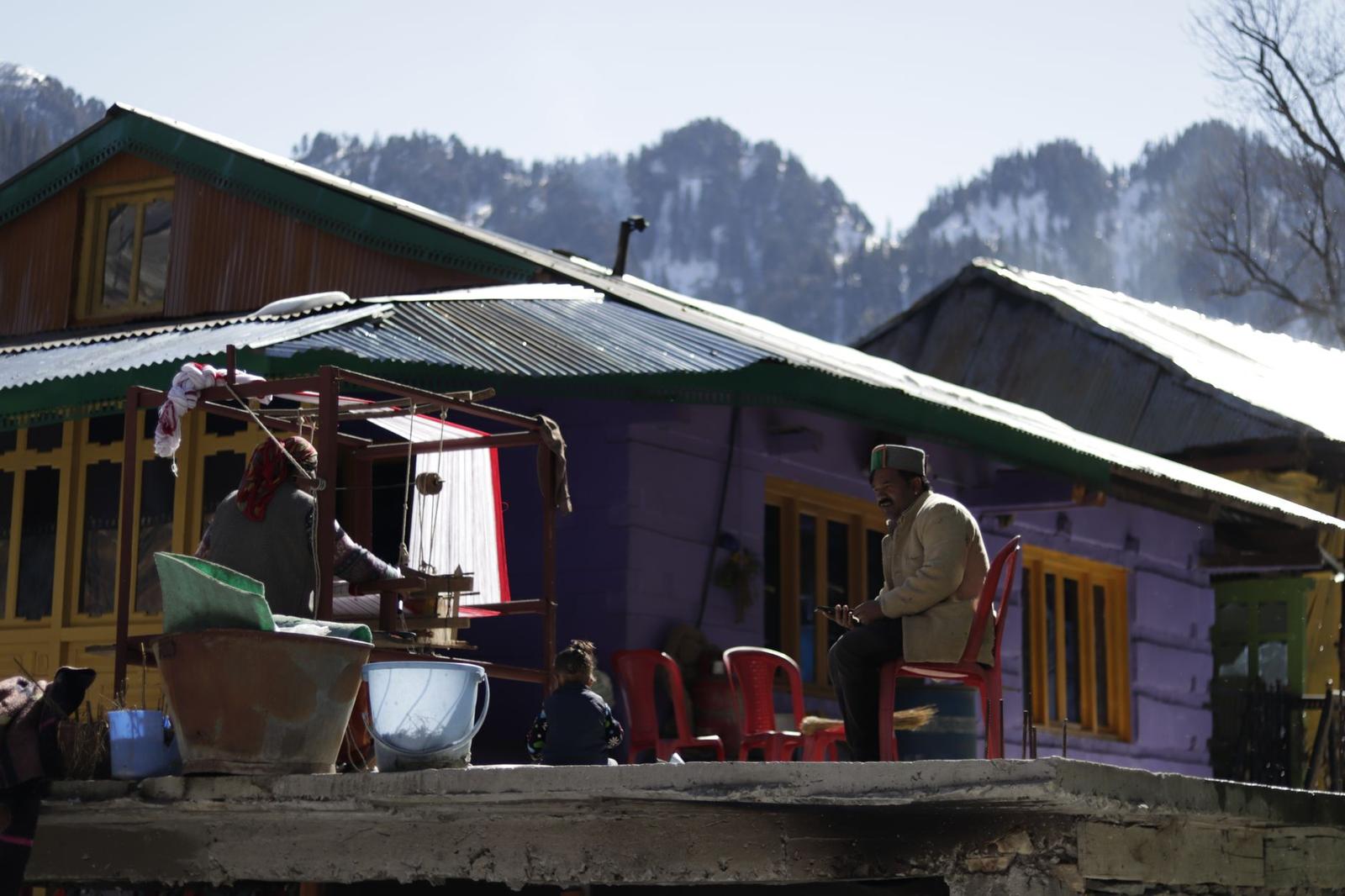A man sitting outside a corrugated-roof homestead beneath snowy peaks