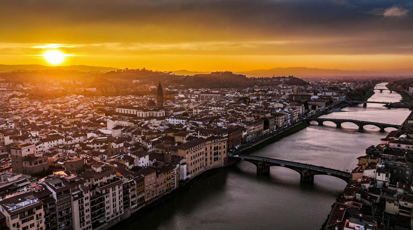 A river city skyline glowing under an orange sunset with stone bridges