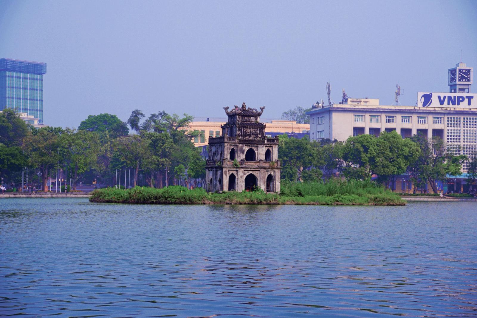 Scenic view of Turtle Tower in Hoan Kiem Lake, Hanoi, framed by greenery and urban backdrop