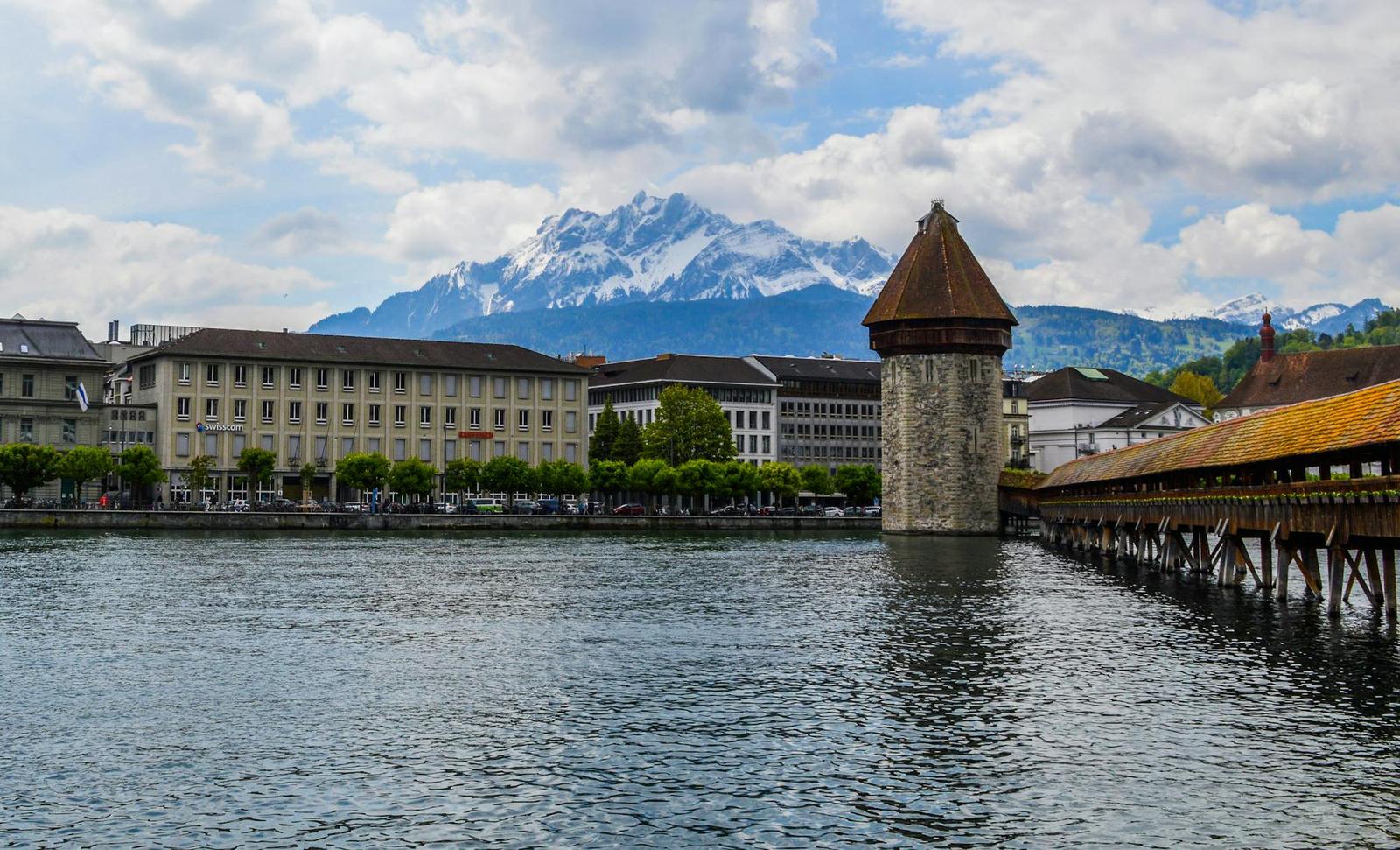 Aged brick tower with footbridge above rippling water of river against snowy mountains in cloudy day