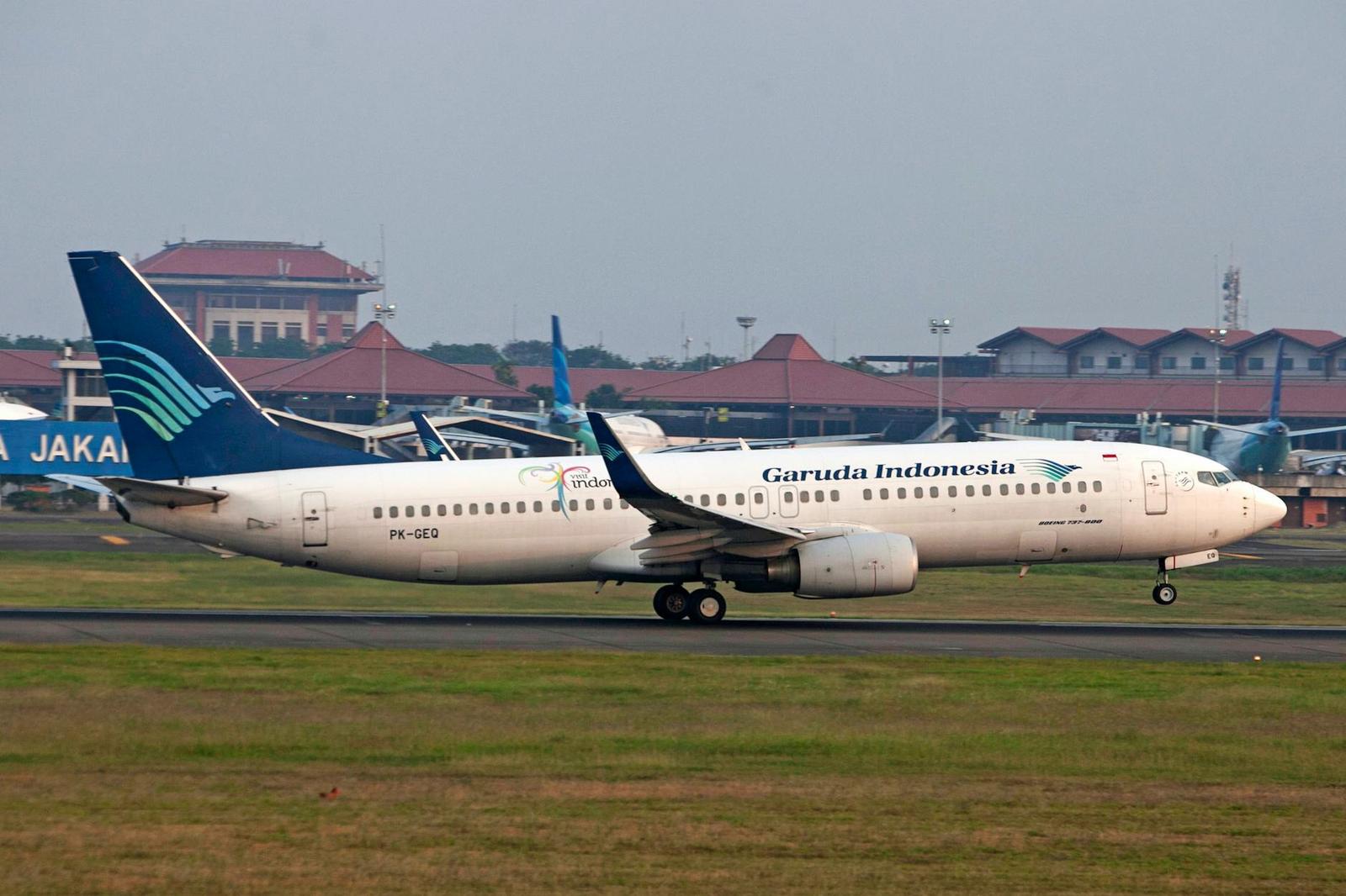 A passenger jet taxis on the runway past red-roofed airport terminal buildings