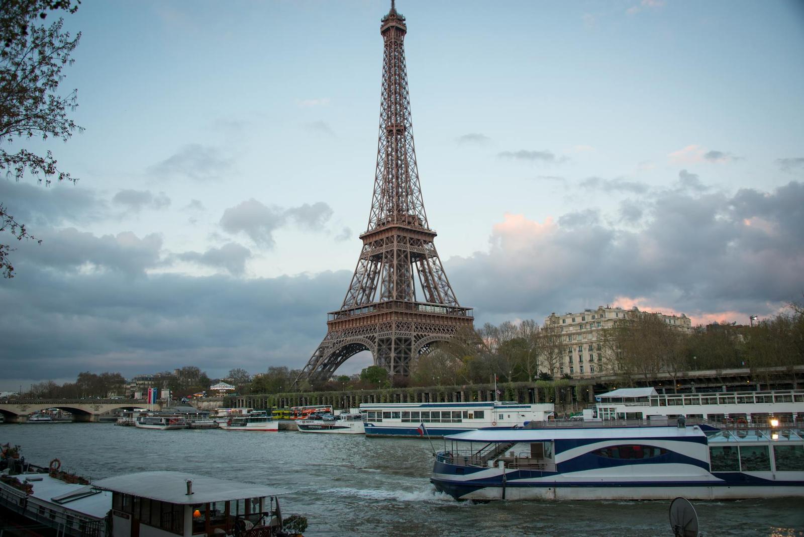 The Eiffel Tower at dusk with tour boats crossing the river in the foreground