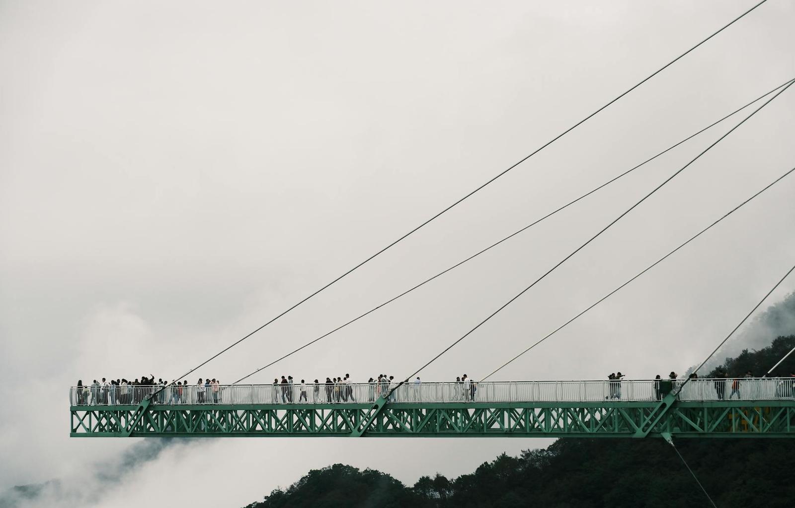 Visitors crossing a green suspension skywalk high above misty forested peaks