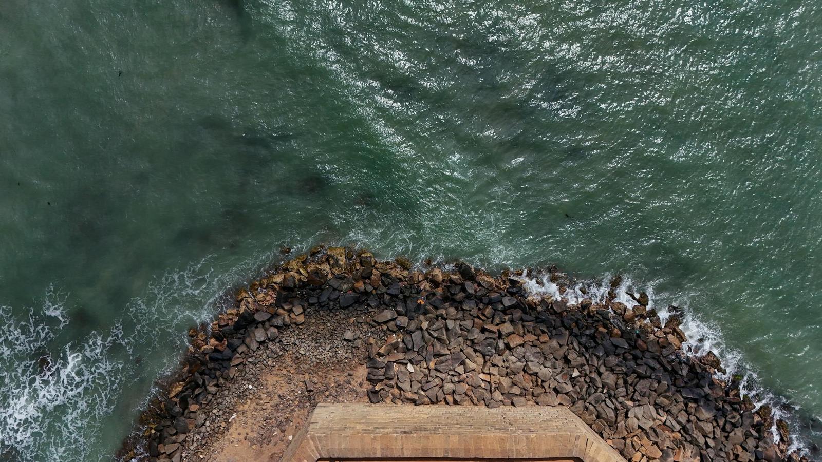 An aerial view of a rocky breakwater jutting into teal waters