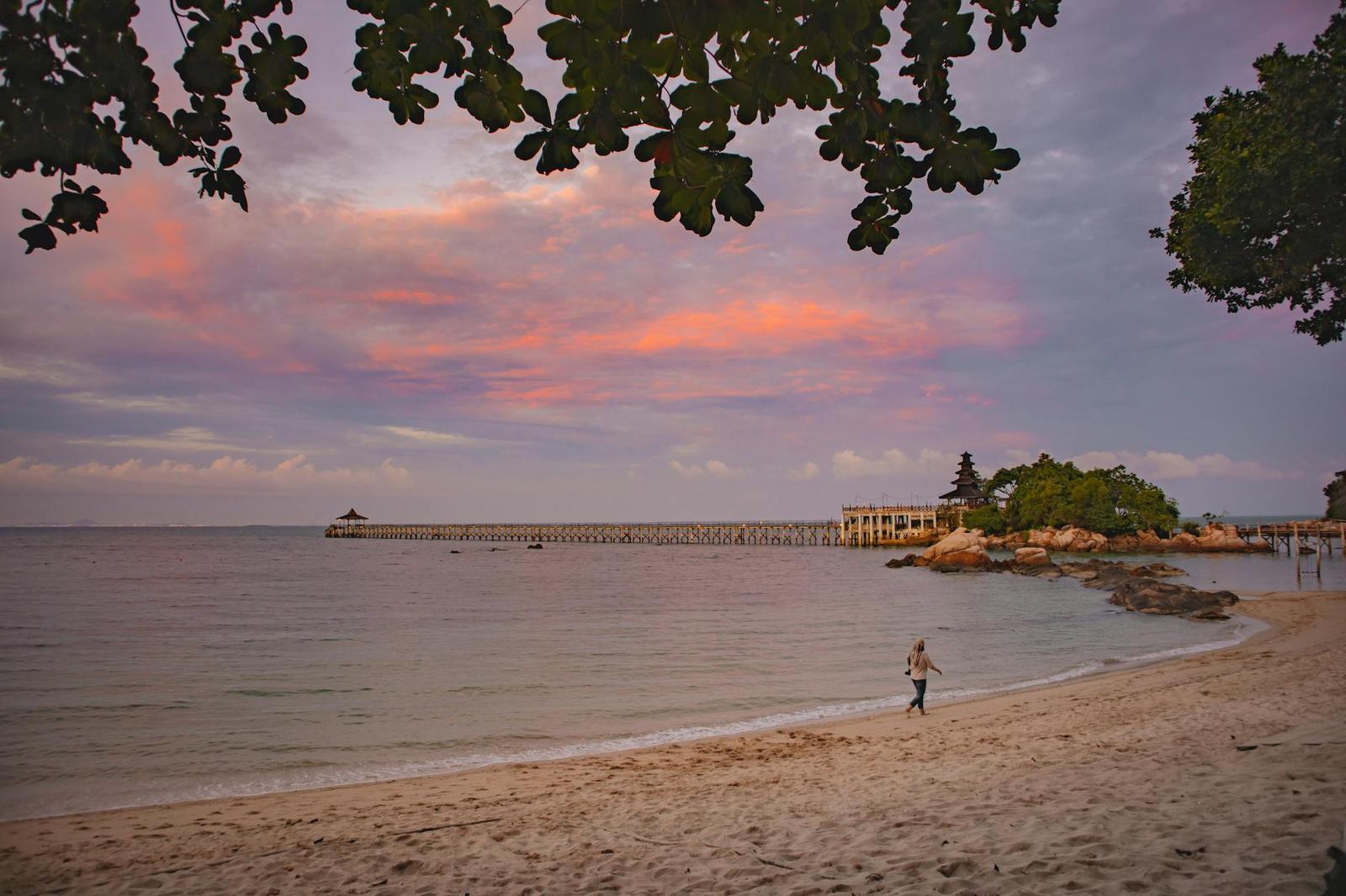 A figure walking along a beach at dusk near a wooden pier and small pavilion