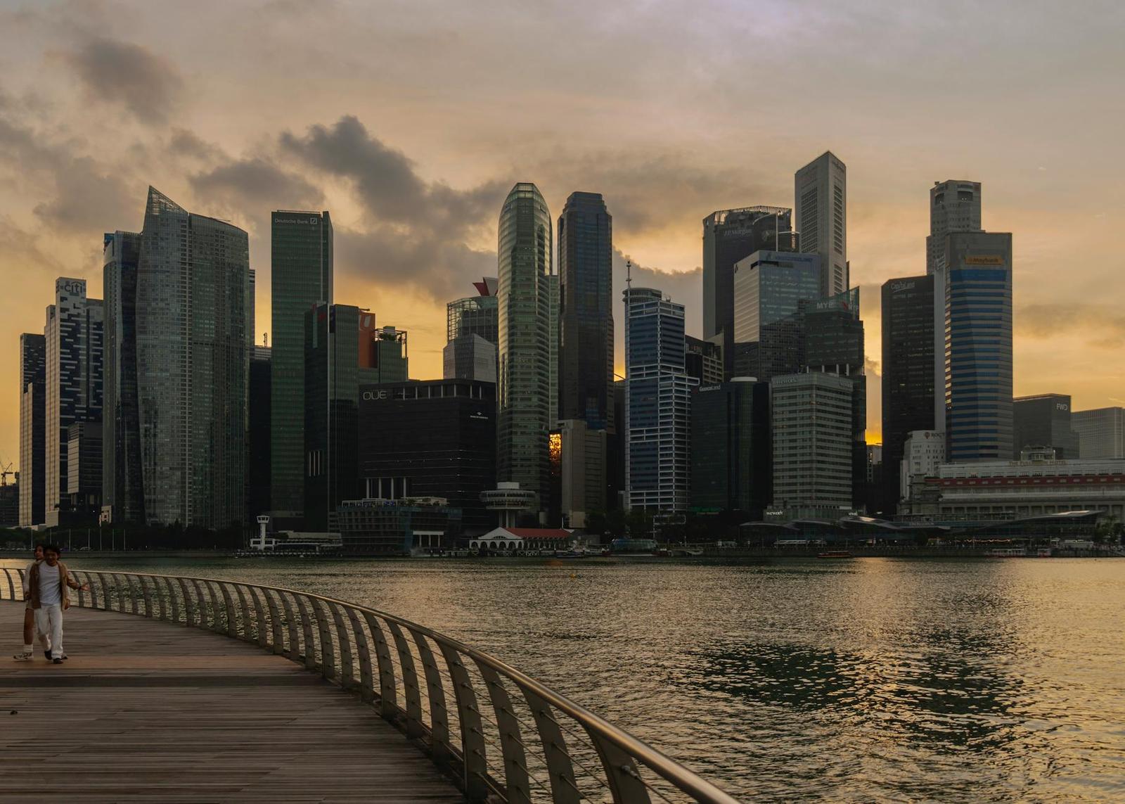 A waterfront promenade with a lone walker facing a glowing city skyline at dusk