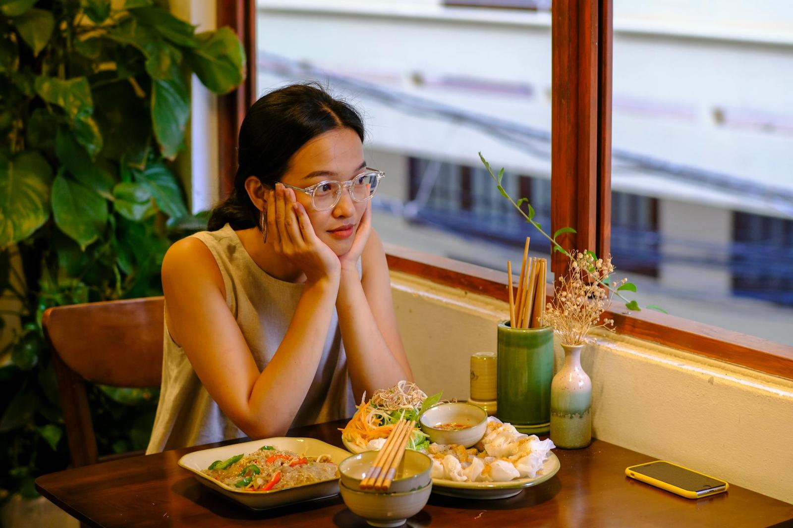 A woman resting her chin on her hands at a table set with noodle dishes by a window