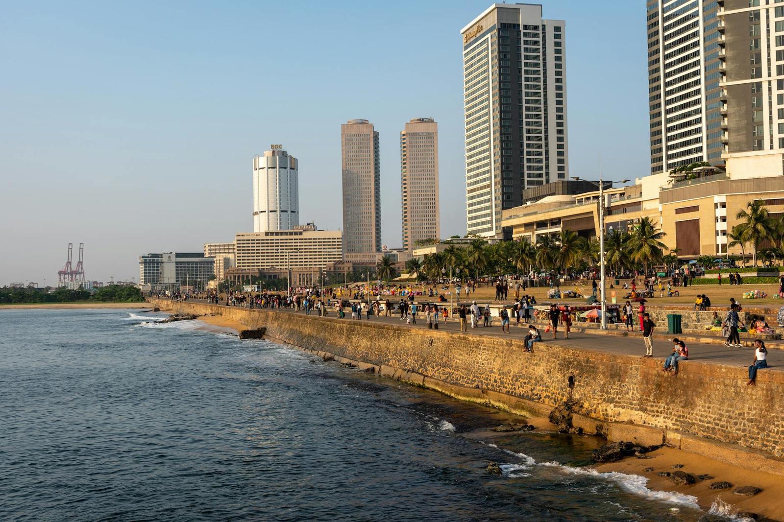Crowds gathered along a seaside stone rampart beneath city towers