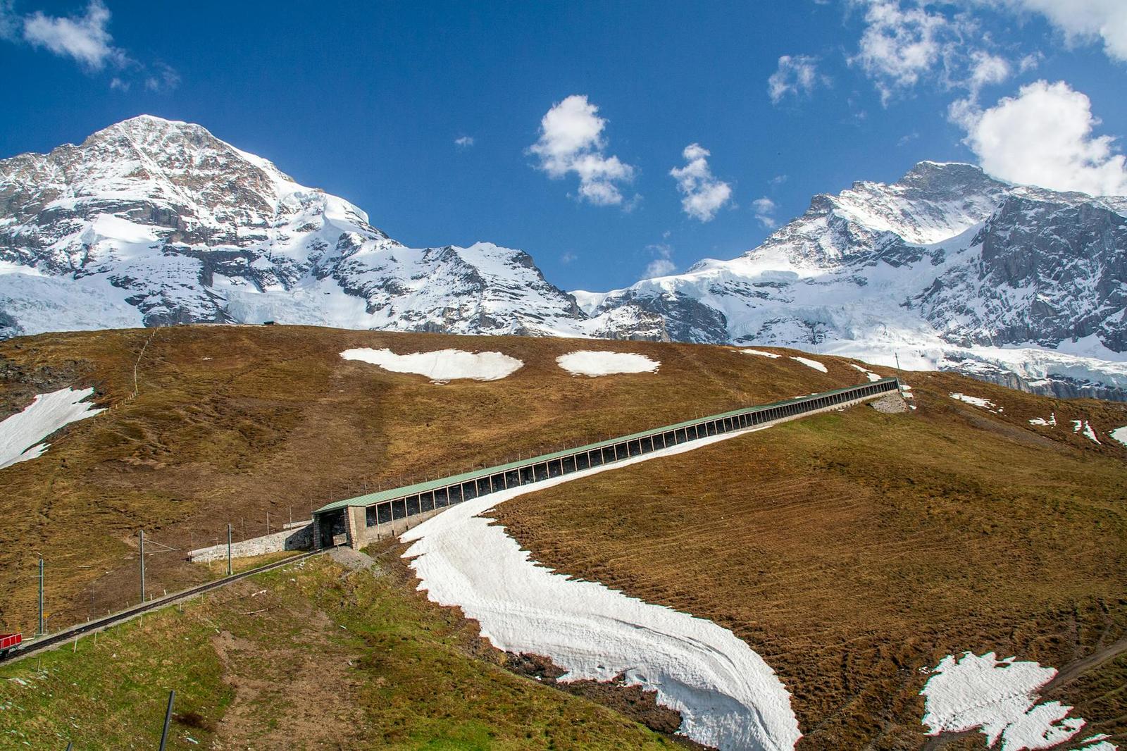 Elevated rail line crossing a snow-patched alpine meadow between peaks