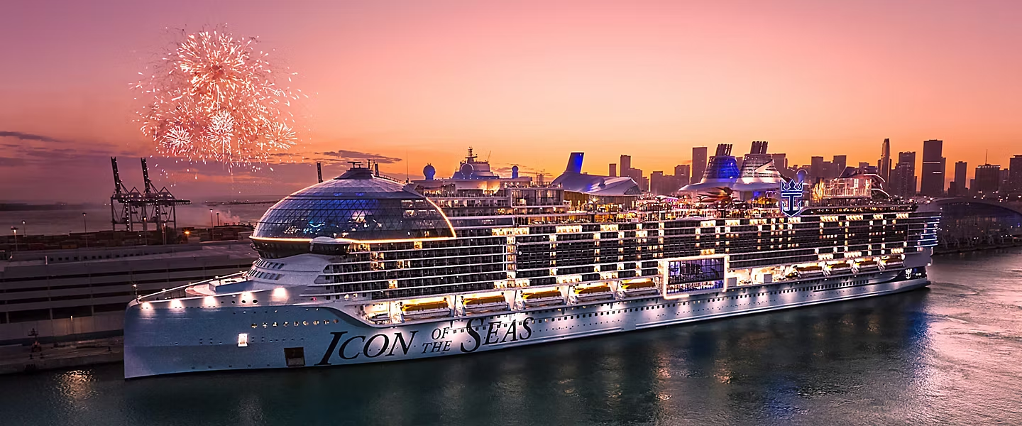 A brightly lit cruise ship docked at sunset with fireworks over the city skyline