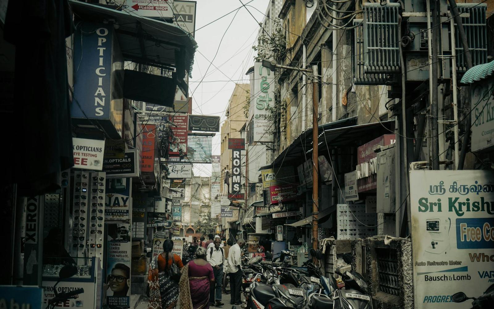 A crowded market street lined with shop signs, wires and parked motorbikes
