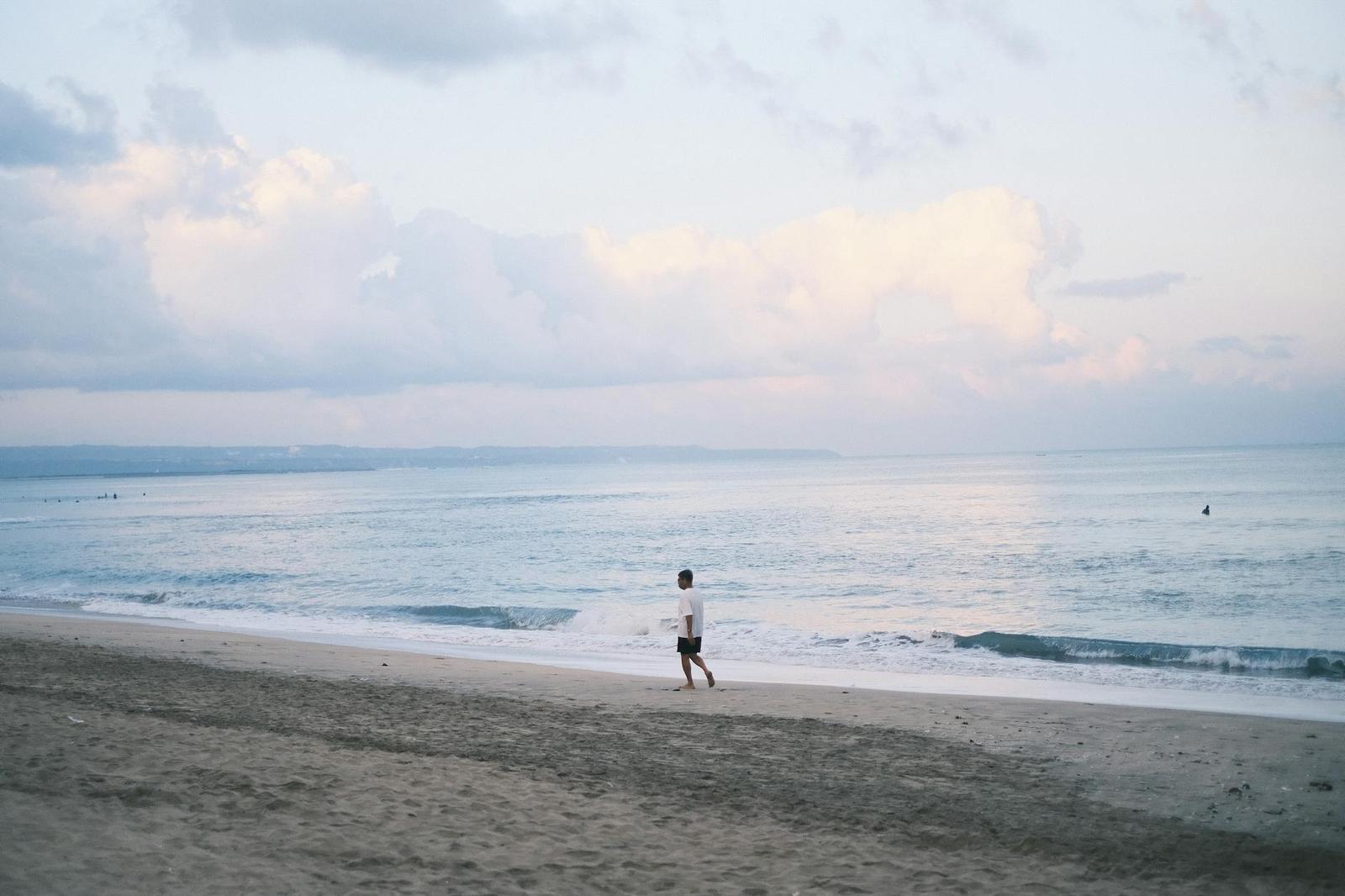 A lone figure walks along a wide sandy beach under overcast sky