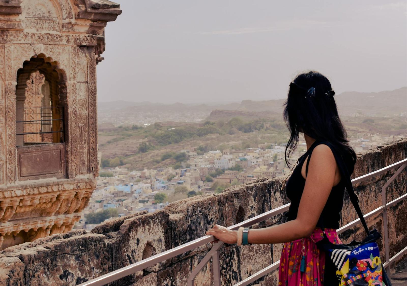 A woman looking out from a hilltop fort balcony over a hazy blue city below