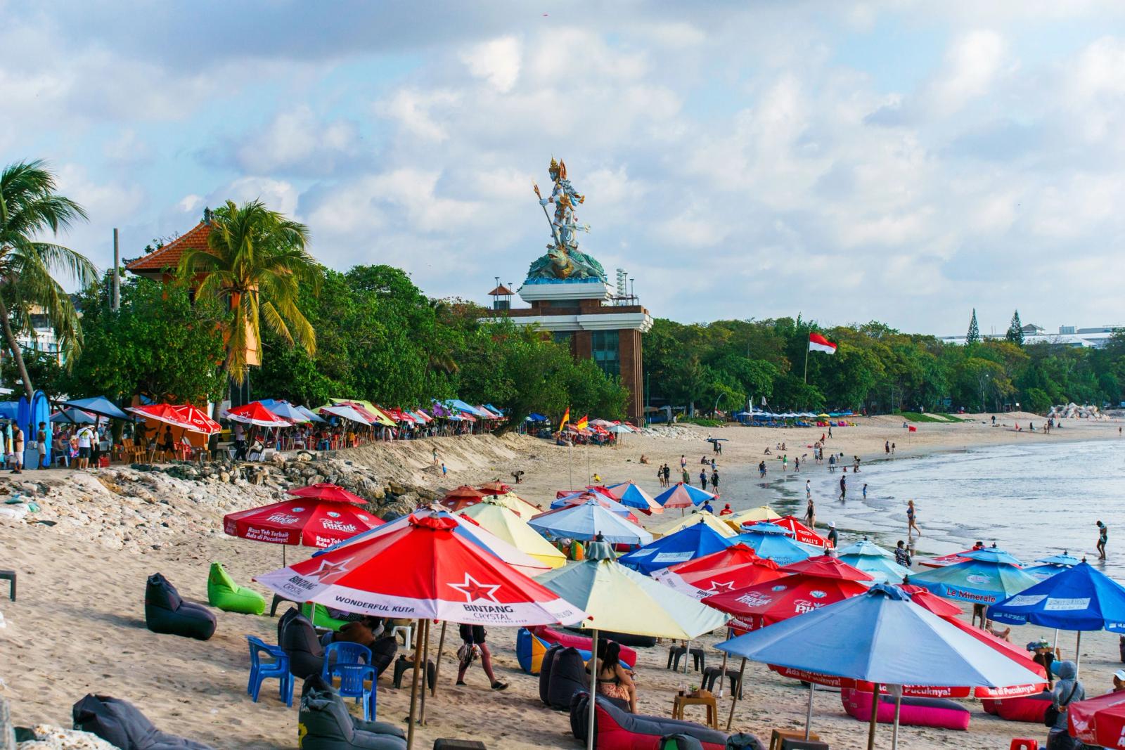 Colorful beach umbrellas and relaxed tourists on a sunny day at Kuta Beach, Bali, with lush greenery in the background