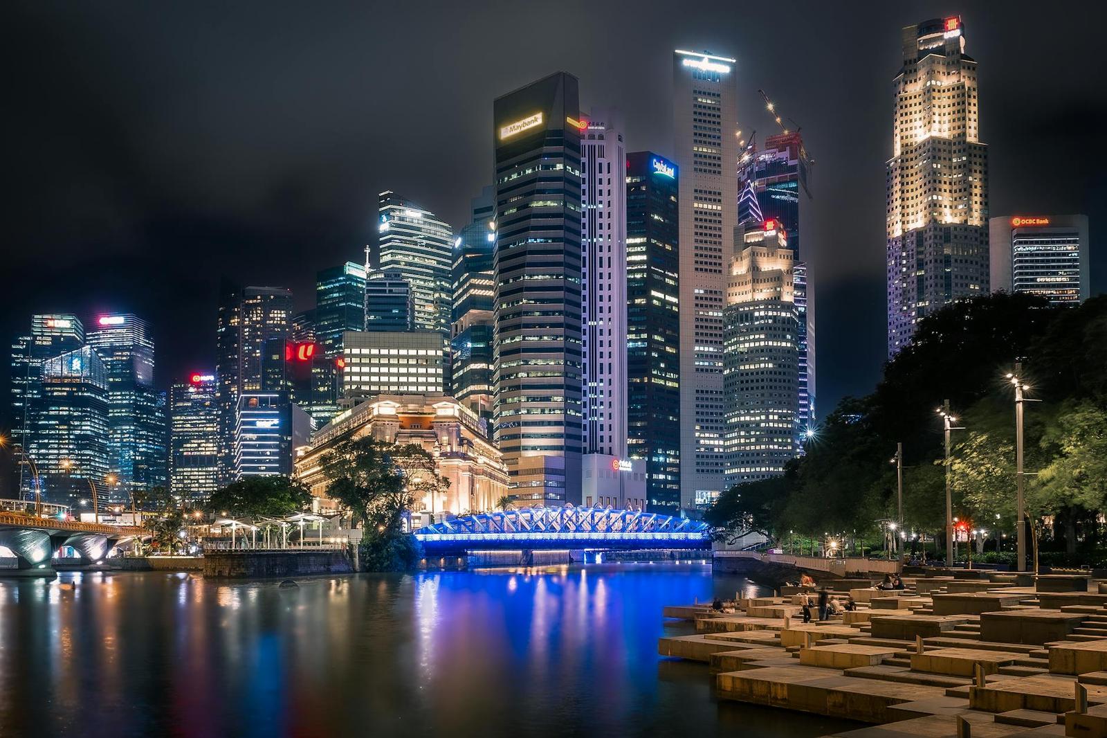 A city skyline glows at night above a riverside promenade with an illuminated bridge